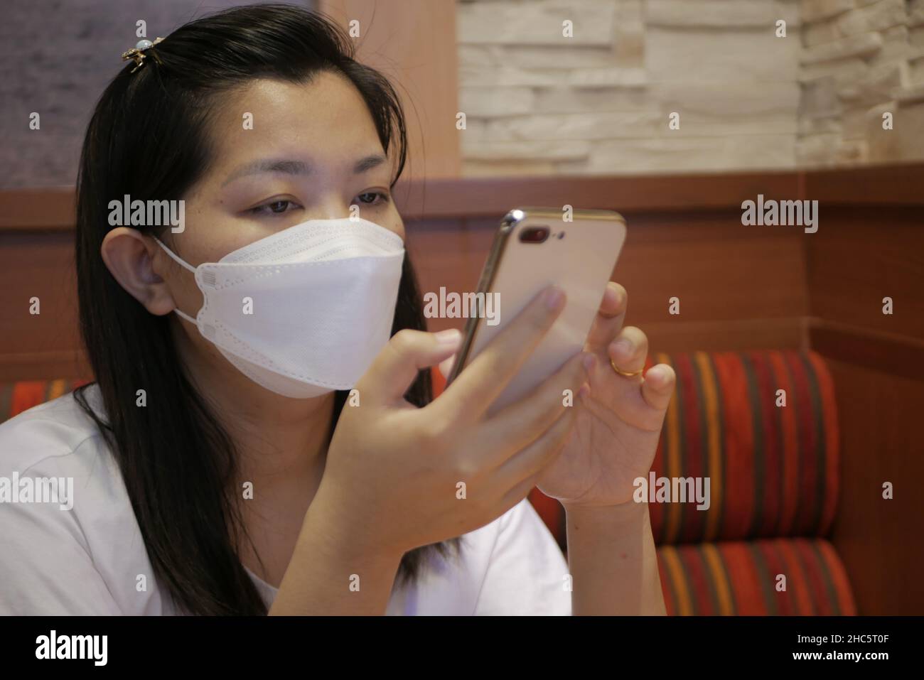 Südostasiatische Frau überprüft ihr Smartphone mit einer medizinischen Maske im Gesicht Stockfoto