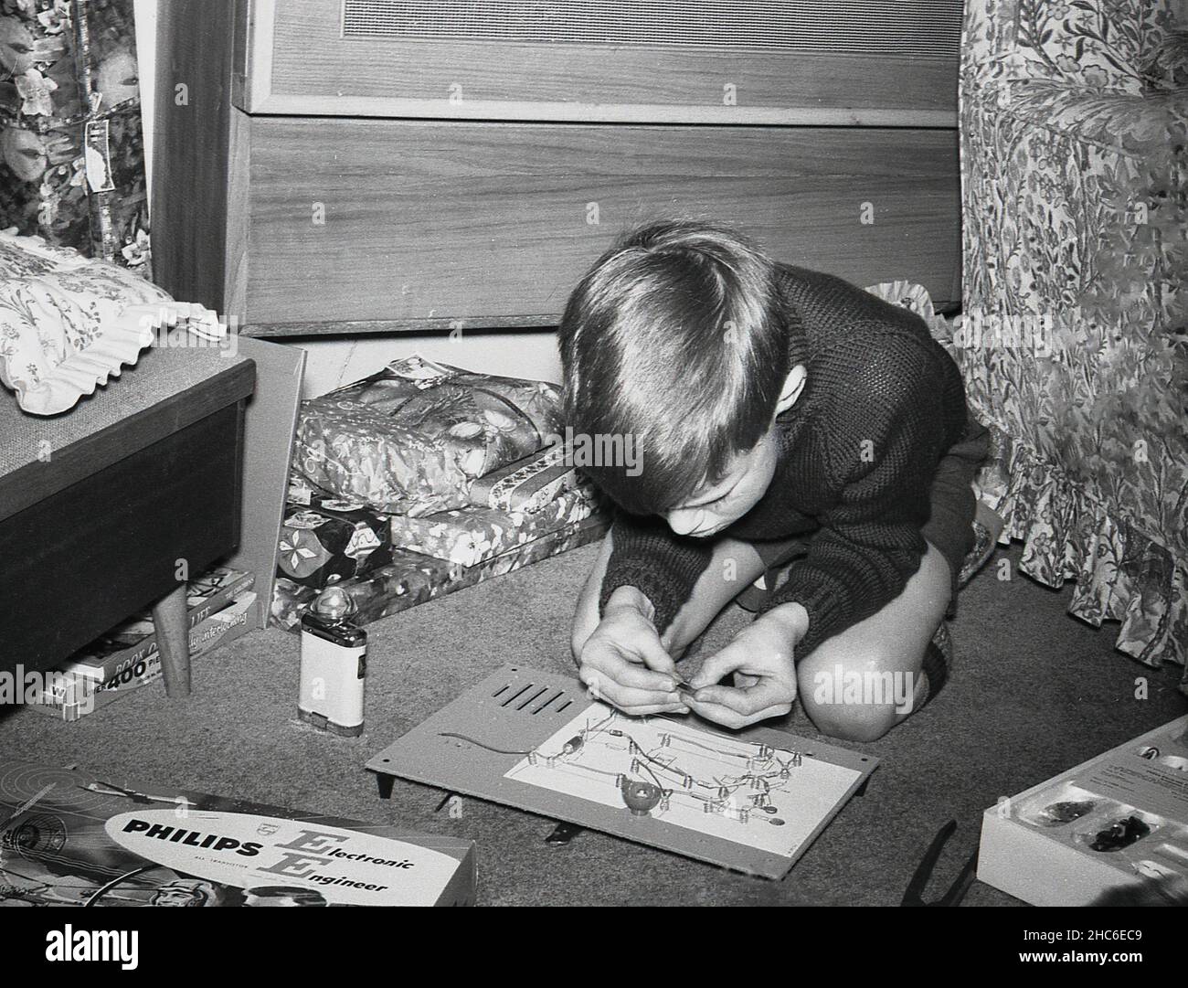 1960s, historisch, in einem Raum, ein kleiner Junge, der auf dem Boden kniet und mit einem weihnachtsgeschenk spielt, eine Platine aus einem Philips Electronic Engineer Kit, England, Großbritannien. Boxed „Young Engineer“-Kits waren eine beliebte pädagogische und unterhaltsame Aktivität für junge Jungen in dieser Zeit. Stockfoto