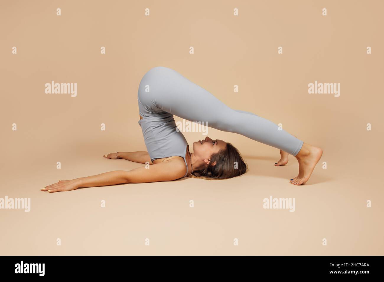 Junge Brünette macht Yoga. Schöne Frau in grau eng anliegender Anzug macht Yoga-Übungen Asana Pflug oder Fitness Stretching auf neutral beige Stockfoto