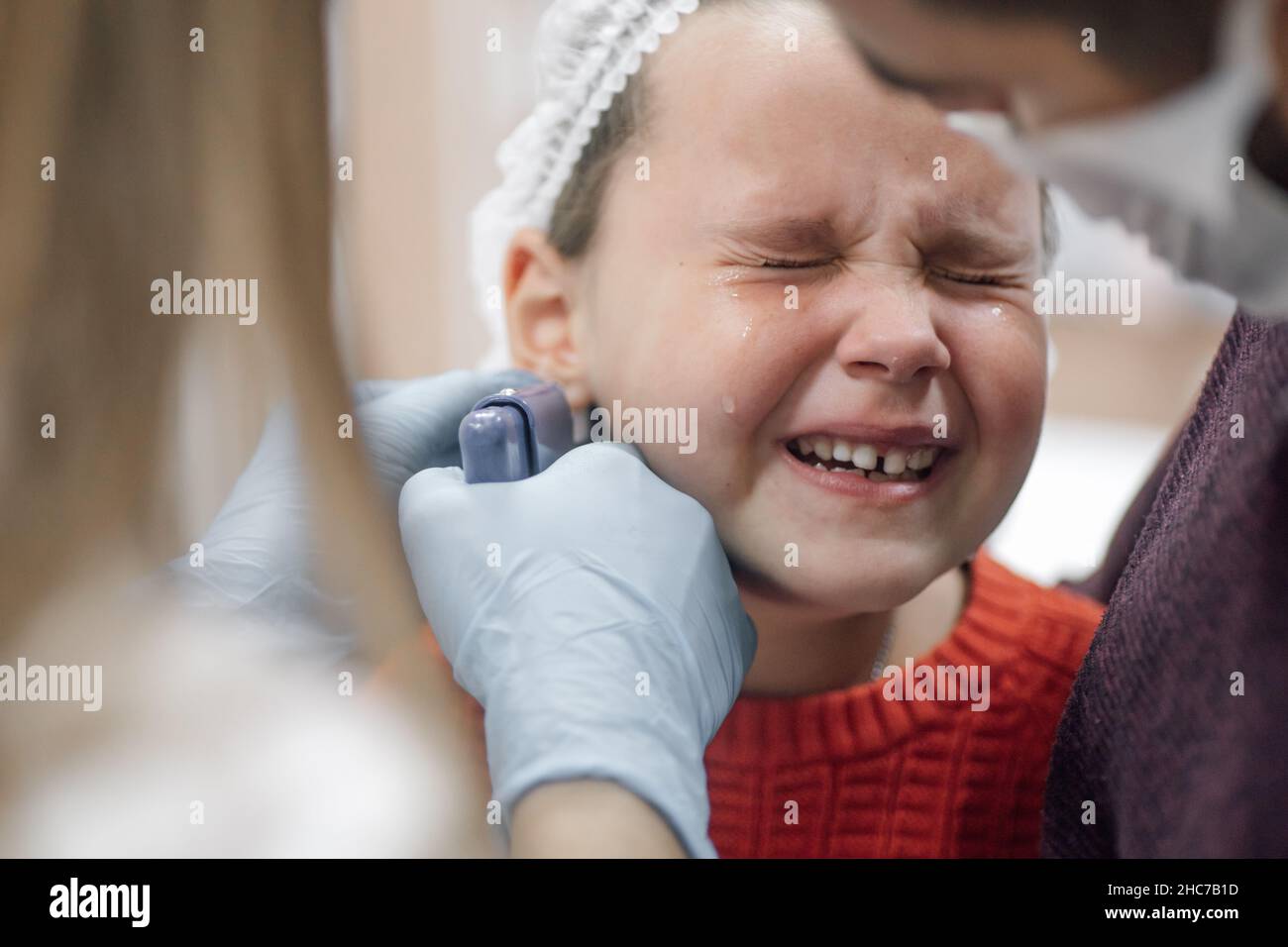 Krankenschwester trägt medizinische Einweg-Handschuhe Durchstechen Ohr mit Pistole für stechende Ohren des Kindes. Kleines Mädchen weint vor Schmerzen. Papa Unterstützung und ruhige Tochter. Stockfoto