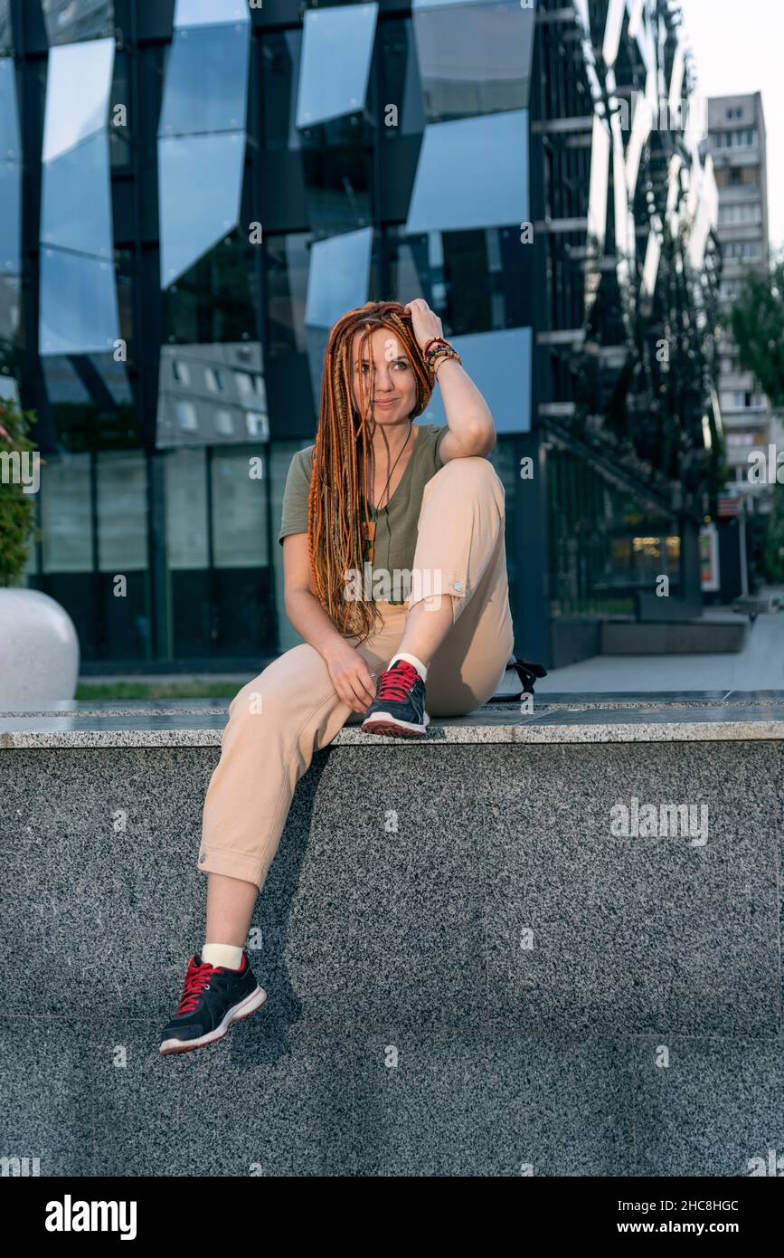 Mädchen mit einem fröhlichen Blick in die Ferne, das auf einer Brüstung in der Nähe eines ungewöhnlichen Bürogebäudes sitzt. Schöner, nicht formeller Büroangestellter. Im Freien Stockfoto