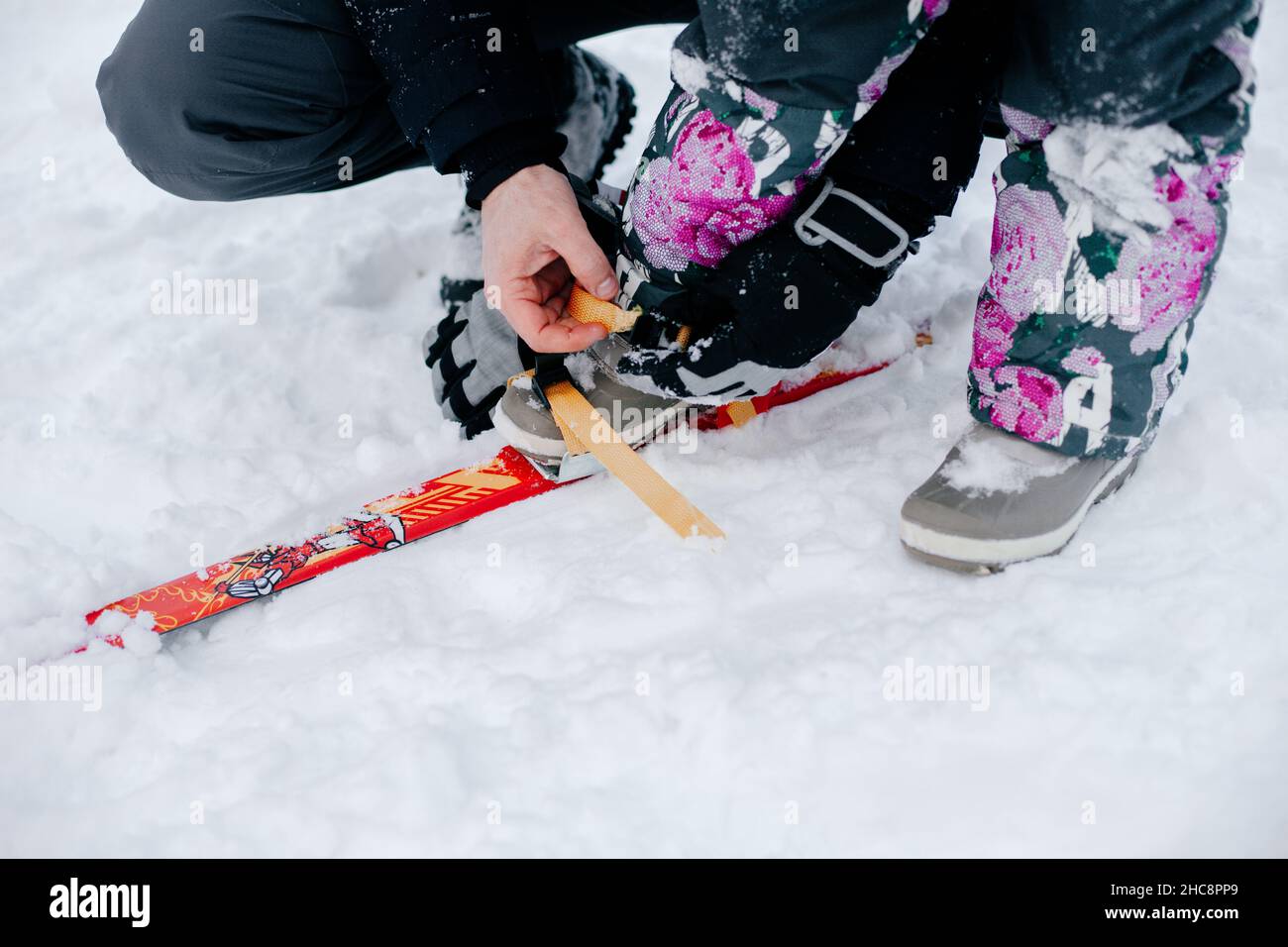 Nahaufnahme eines weiblichen Kindes, dem der liebevolle Vater dabei hilft, Ski auf die Füße des Kindes zu setzen, ohne Gesichter im Rahmen zu haben. Erstaunlicher Hintergrund voller Weiß Stockfoto