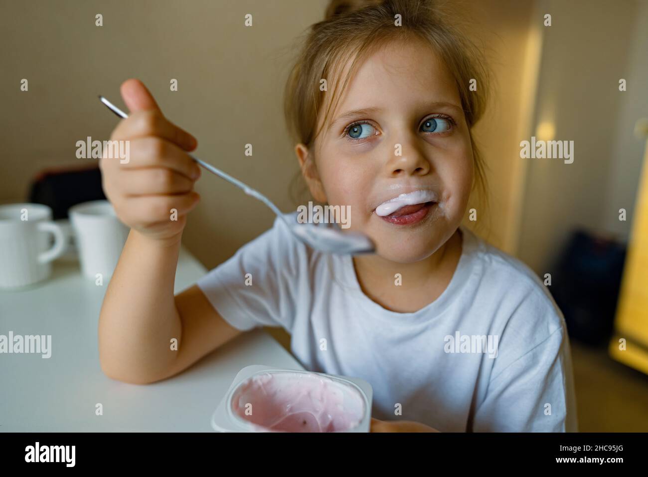 Nettes Mädchen isst Joghurt mit Appetit, hat schmutzigen Mund, hat Spaß, Grimassen Stockfoto