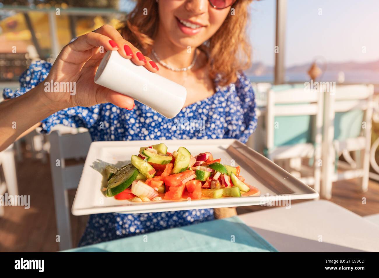 Glückliche Frau mit Salzkeller in der Hand würzen Salat mit überschüssigem Salz oder Pfeffer in einem Restaurant. Ungesunde Essgewohnheiten führen zu Krankheiten Stockfoto