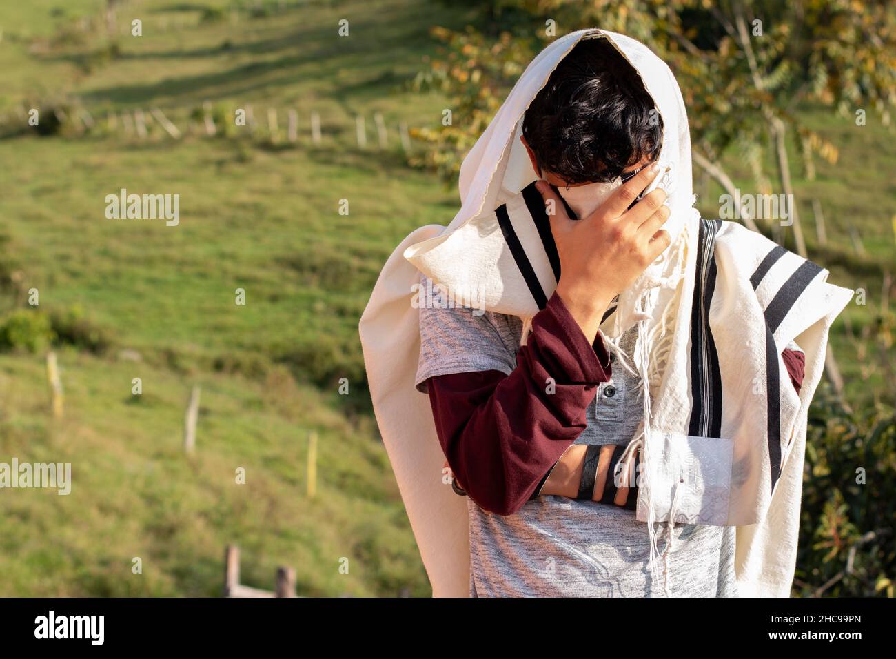 Jude trägt Tallit und Tefillin oder Mylakterie und betet in der Natur, während er sein Gesicht mit den Händen bedeckt. Stockfoto