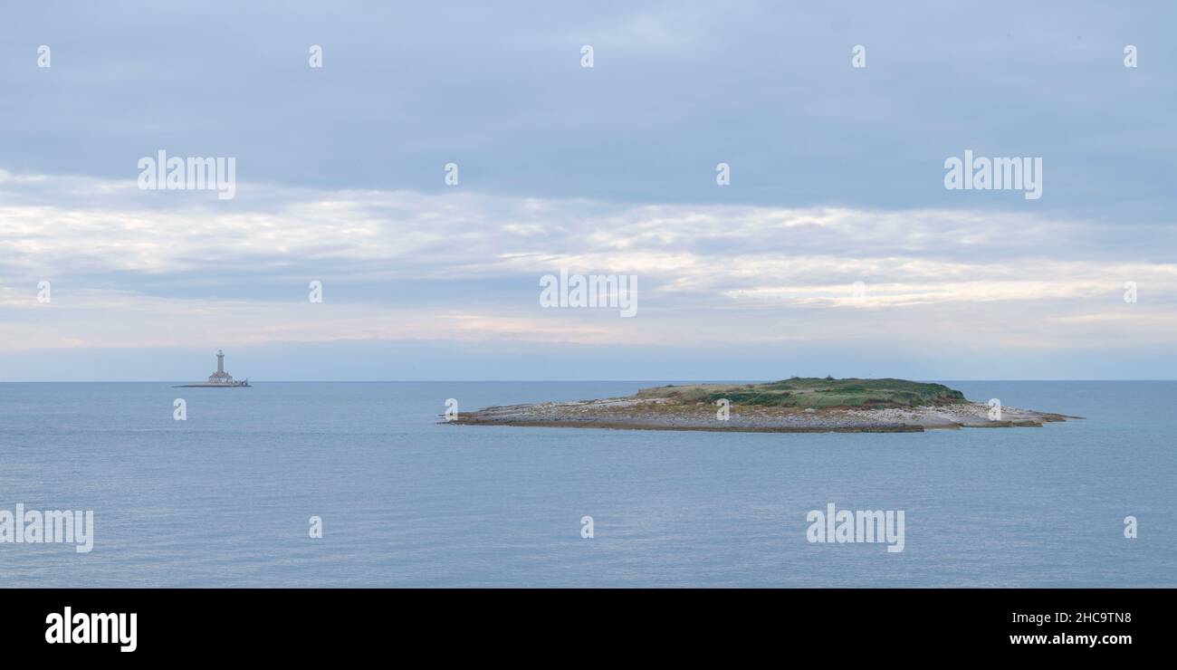 Kleine felsige Inseln mit felsiger Küste im Vordergrund und Leuchtturm im Hintergrund, nebliger Atmosphäre und bewölktem Himmel Stockfoto