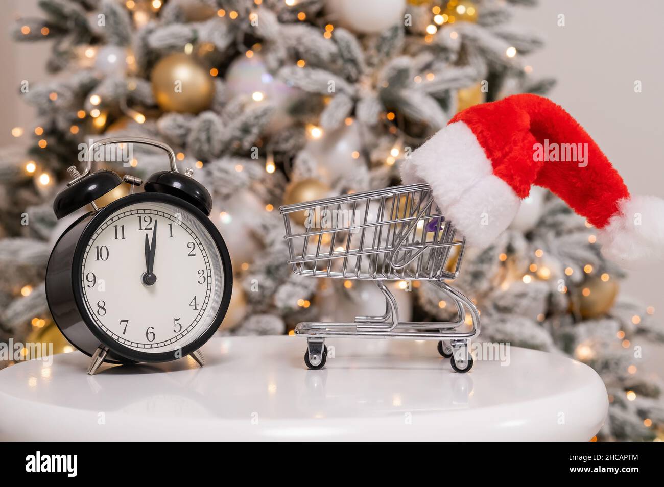 Weihnachtsgeschenke Zeit zum Einkaufen. Ein Einkaufswagen mit Weihnachtsmütze und Wecker am Weihnachtsbaum. Stockfoto