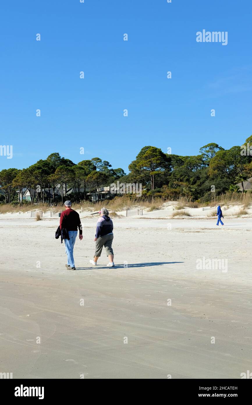 Rückansicht eines älteren Paares, das am Strand in Hilton Head, South Carolina, spazieren geht; an einem sonnigen Tag mit Bäumen im Hintergrund einen Spaziergang im Sand macht. Stockfoto