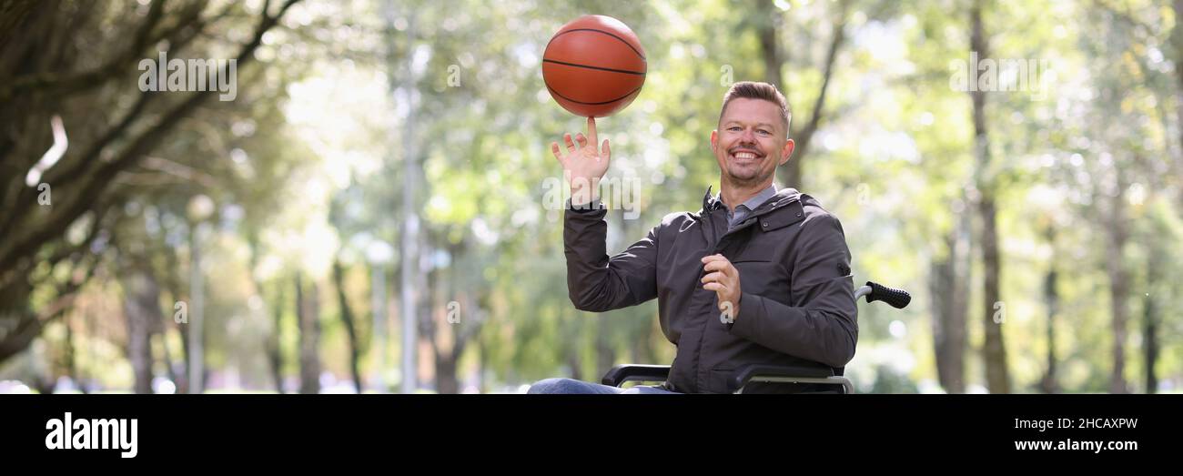 Lächelnder Mann dreht Basketball auf dem Finger, während er im Rollstuhl im Park sitzt Stockfoto