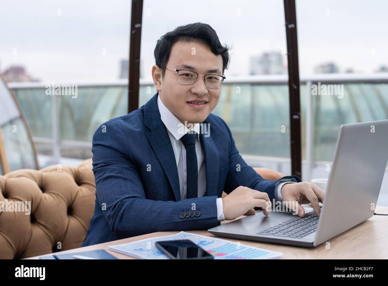 Fröhlicher junger chinesischer Geschäftsmann in formeller Kleidung und Brillen, der vor dem Laptop arbeitet, während er im Café am Tisch sitzt Stockfoto