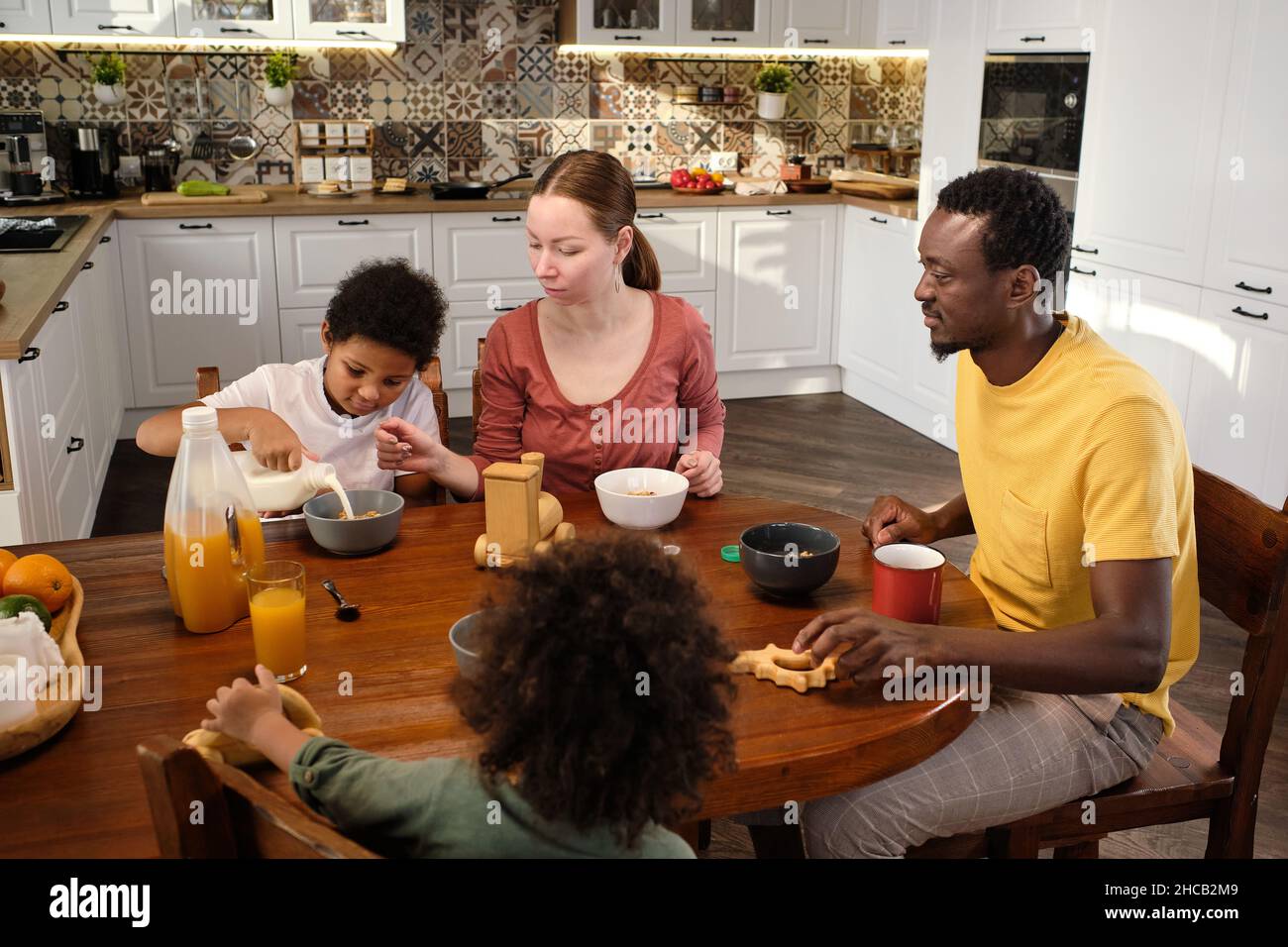 Moderne vierköpfige interrassische Familie, die beim Frühstück am hölzernen Küchentisch sitzt Stockfoto