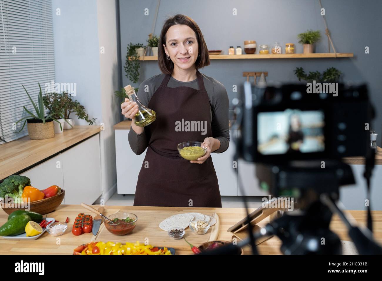 Junge Frau mit einer Flasche Olivenöl und einer Schüssel mit Gewürzen, die während des Masterclasses für das Online-Publikum am Küchentisch steht Stockfoto