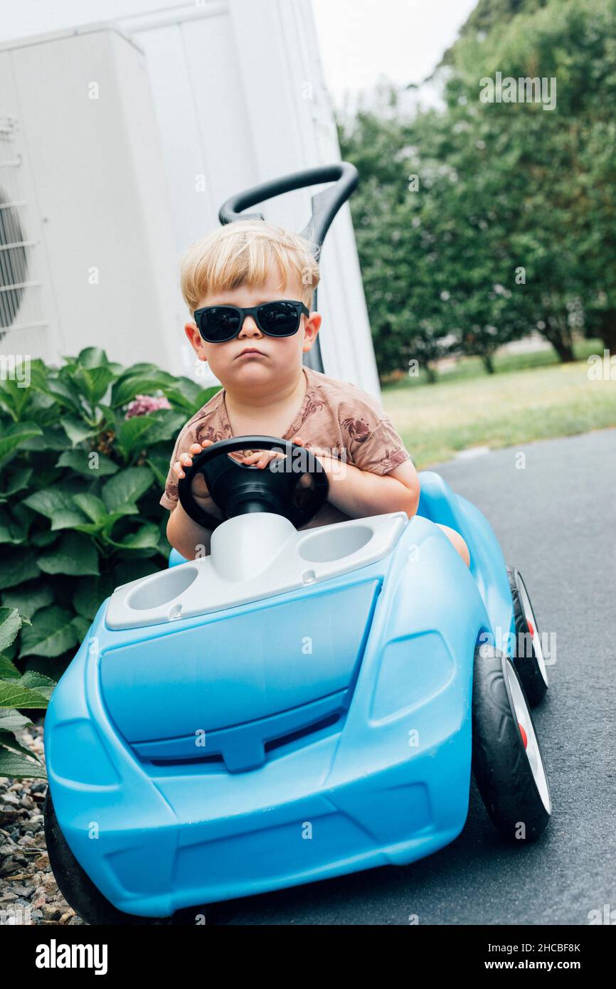 Gelangweilter blonder Junge mit Sonnenbrille im blauen Spielzeugauto Stockfoto