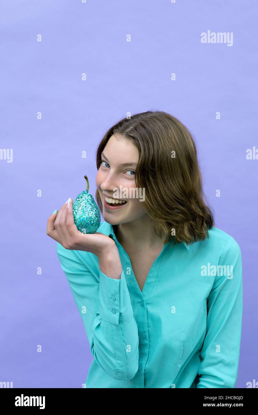 Lächelndes Teenager-Mädchen hält Birne vor dem Mund vor Lavendel Hintergrund Stockfoto