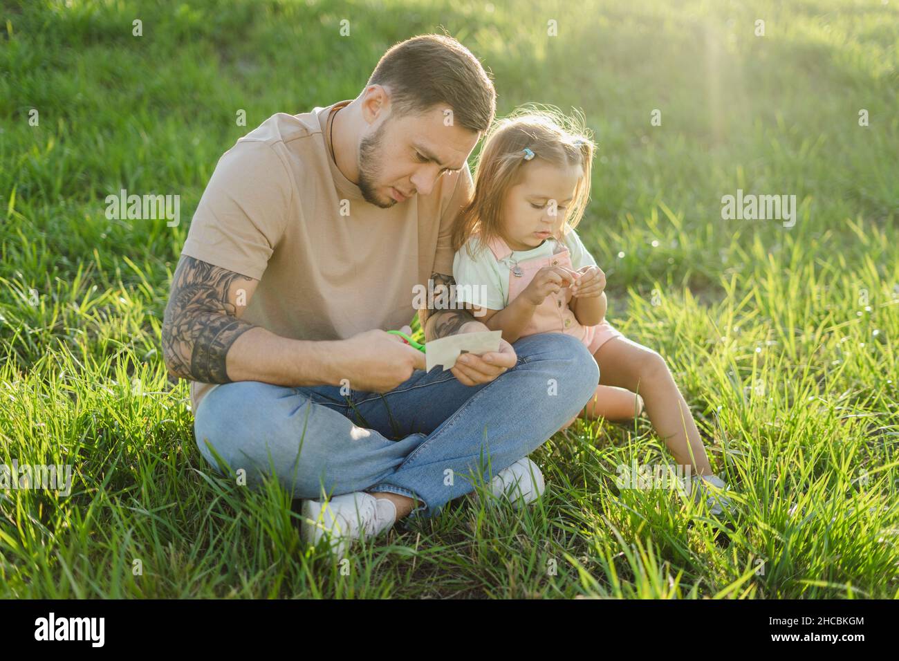 Mann, der Tattoos schneidet, sitzt mit gekreuzten Beinen von der Tochter auf dem Gras Stockfoto