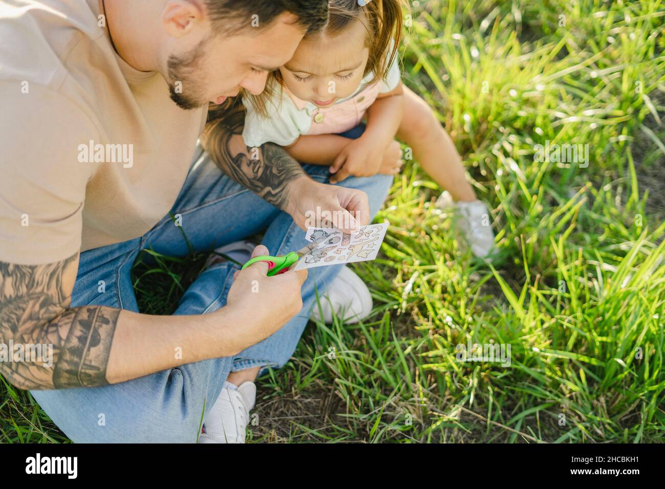 Neugieriges Mädchen, das von einem Vater sitzt und übertragbare Tattoos auf Gras schneidet Stockfoto