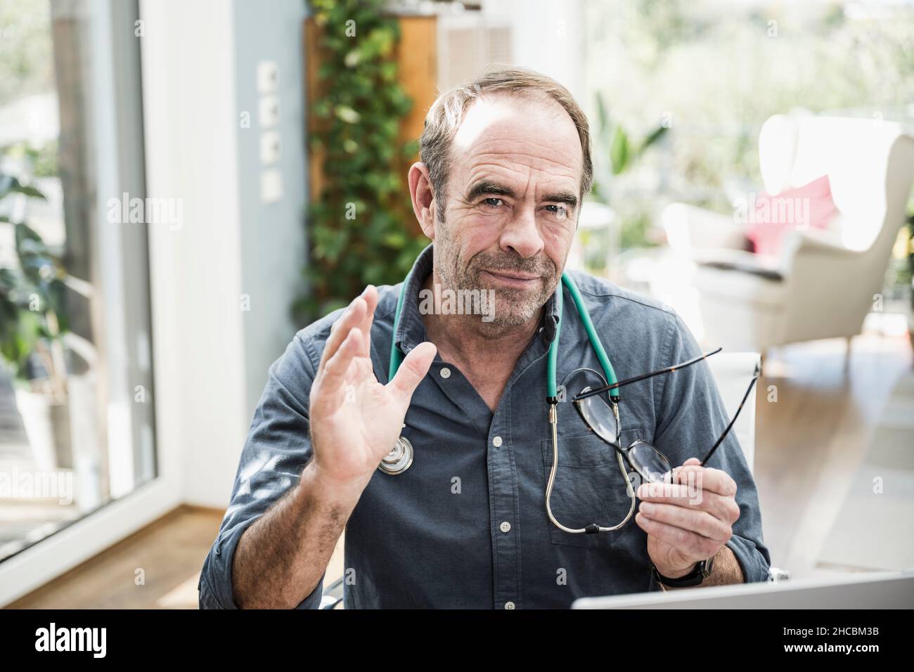Arzt mit Haarstoppel, der eine Brille hält, die in der Klinik winkt Stockfoto
