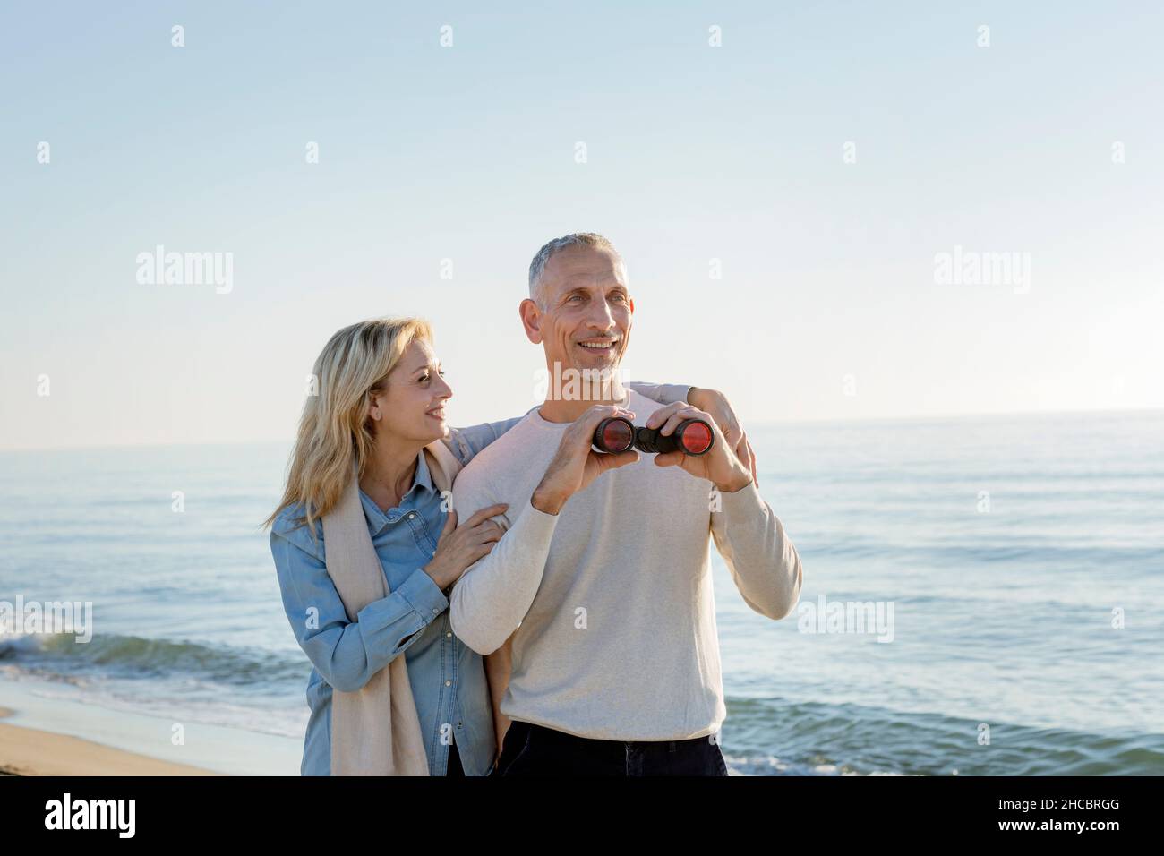 Lächelnde Frau mit Arm um den Mann, der ein Fernglas am Strand hält Stockfoto