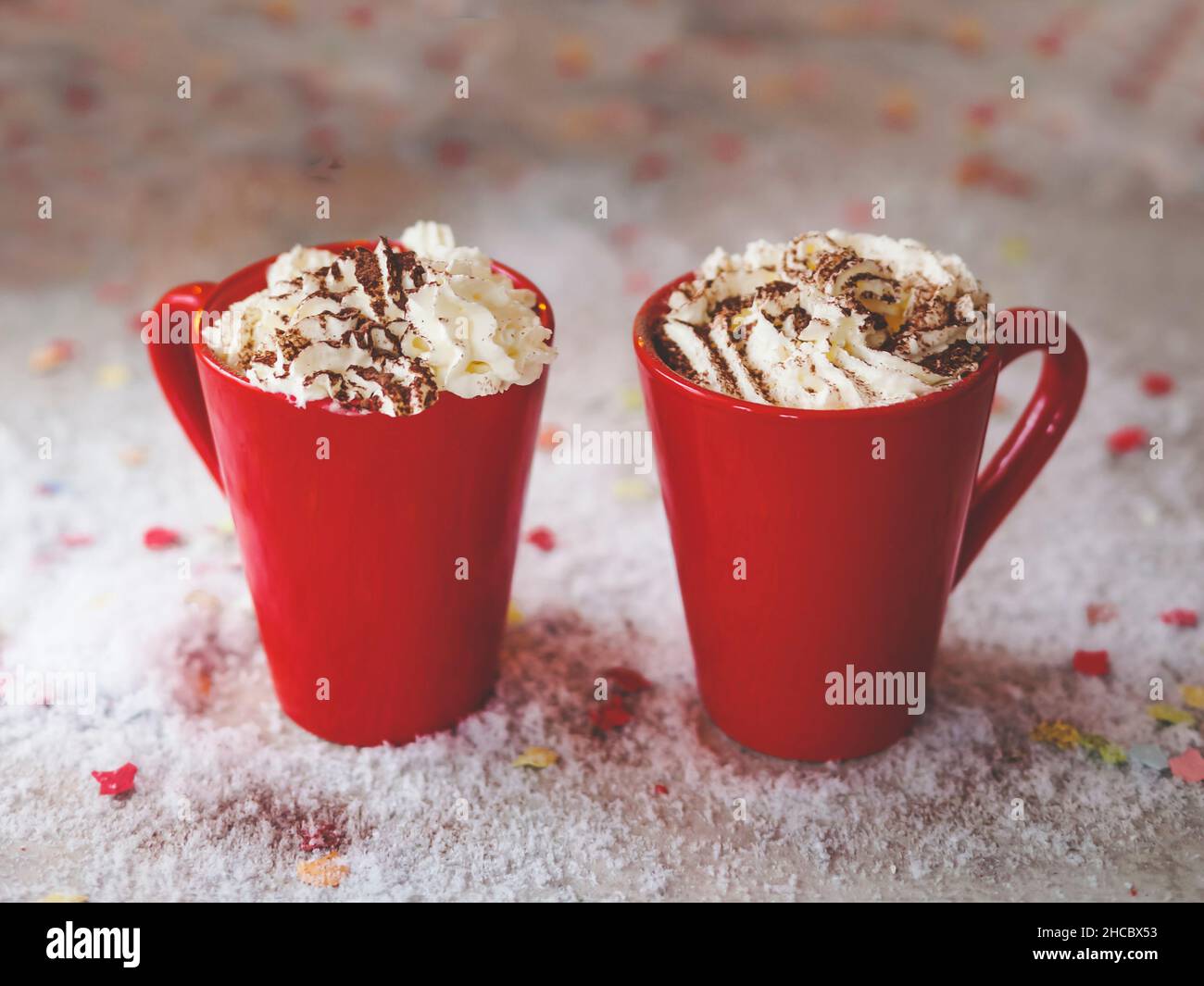 Zwei rote Becher mit heißer Schokolade und Schlagsahne auf dem Tisch mit Schnee. Gemütliche warme Getränke zu Hause mit süßer Creme. Vorderansicht. Stockfoto