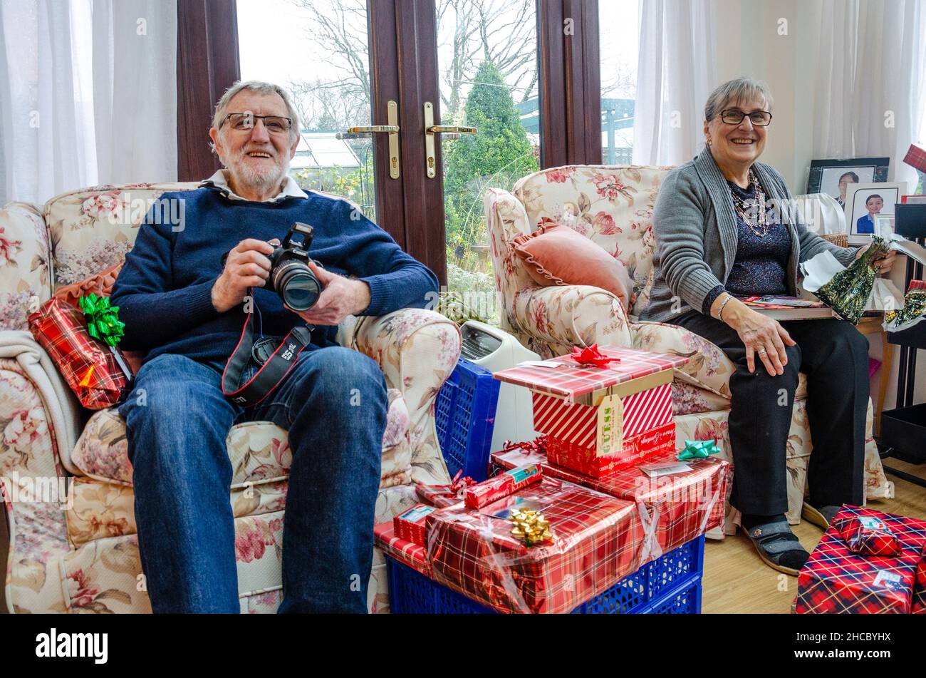 Ein Rentnerpaar sitzt am Weihnachtsmorgen in Sesseln mit Geschenken zum Öffnen, aber schaut glücklich zu, wie die Familie unsere von geschossenen offenen Geschenken zuerst schaut. Stockfoto