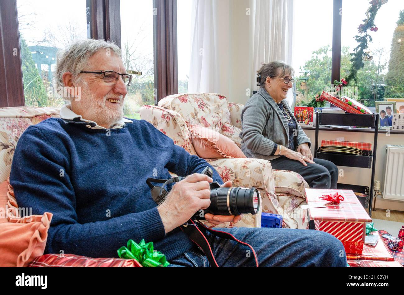 Ein Rentnerpaar sitzt am Weihnachtsmorgen in Sesseln mit Geschenken zum Öffnen, aber schaut glücklich zu, wie die Familie unsere von geschossenen offenen Geschenken zuerst schaut. Stockfoto
