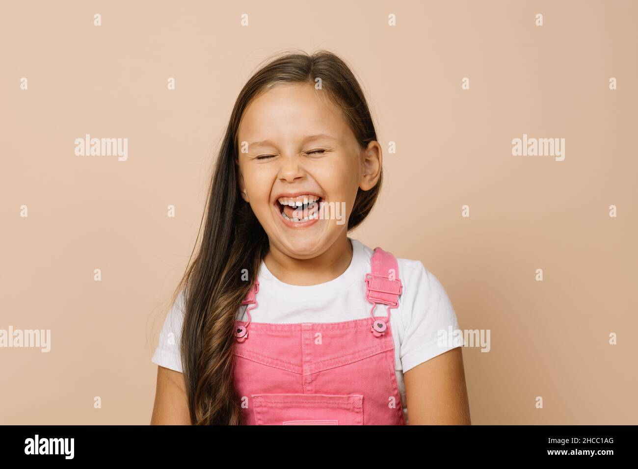 Porträt eines Kindes mit vollständig geöffnetem Mund, geschlossenen Augen und aufgeregt Lächeln mit Zähnen Blick auf die Kamera in leuchtend rosa Jumpsuit und weißem T-Shirt Stockfoto