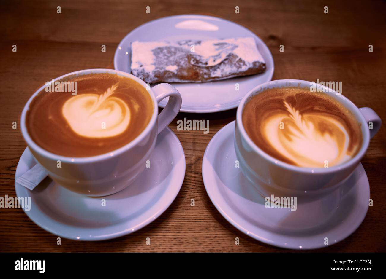 Zwei Cappuccino-Tassen aus weißer Keramik und Kirschkuchen auf einem weißen Keramikteller mit einem Holztisch im Hintergrund Stockfoto