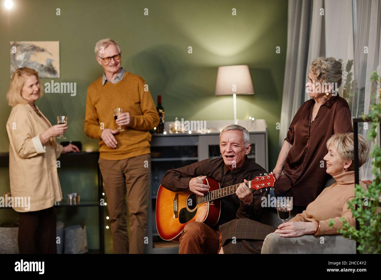 Talentierter Mann im Alter von Kaukasien, der beim Treffen mit Freunden akustische Gitarre spielt und singt. Vier Senioren lauschen ihrer gebratenen Darbietung Stockfoto