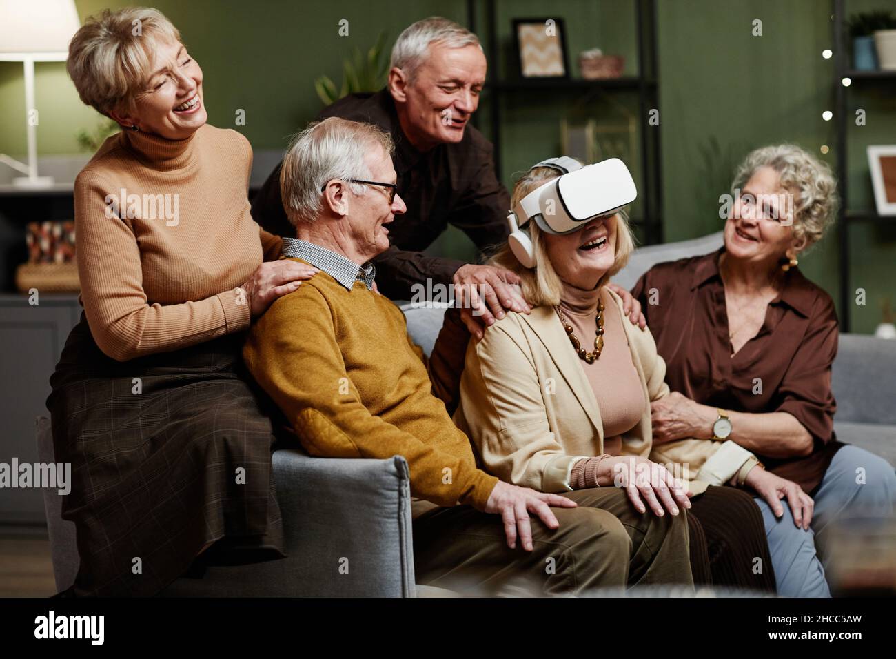 Ältere kaukasische Frau, die auf dem Sofa saß und moderne VR-Brillen genoss, während sich ihre vier Freunde um sie versammelten und sie beobachteten Stockfoto
