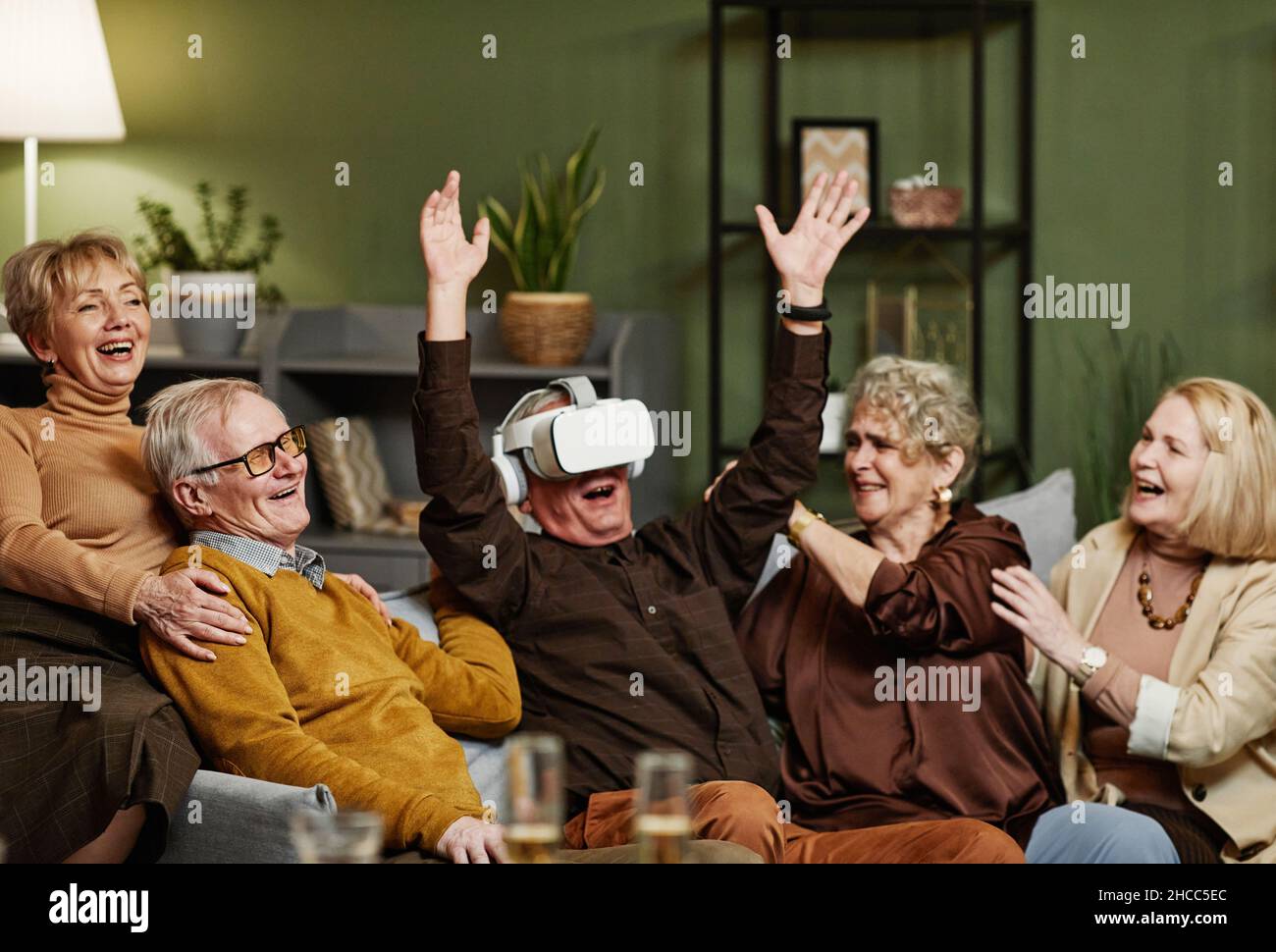 Fröhlicher alter kaukasischer Mann, der auf der Couch saß und moderne VR-Brillen genoss, während sich seine vier Freunde um ihn versammelten und sie beobachteten Stockfoto