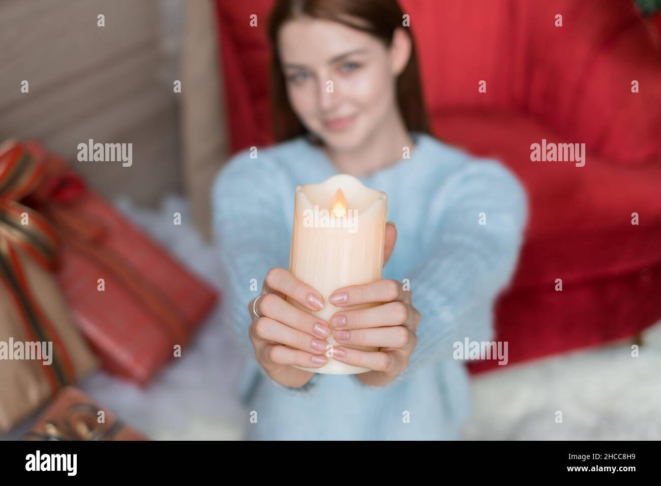 Entzückendes Mädchen sitzt und hält dicke weiße Wachskerze in ihren Händen. Verschwommener Hintergrund, Nahaufnahme, Weihnachtsfeiertage. Stockfoto