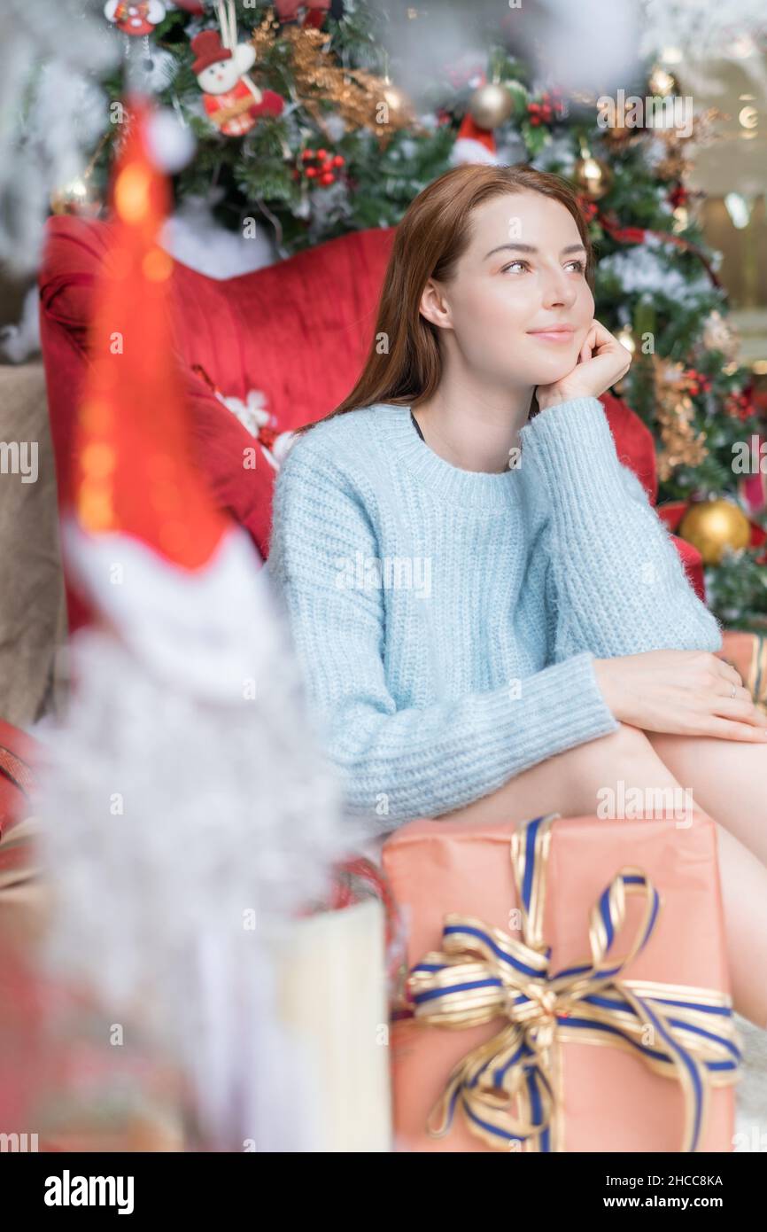Neujahrs-Porträt der schönen kaukasischen jungen Frau in gemütlichen hellblauen Pullover sitzt neben geschmückten Weihnachtsbaum mit Geschenkboxen mit Schleifen Stockfoto