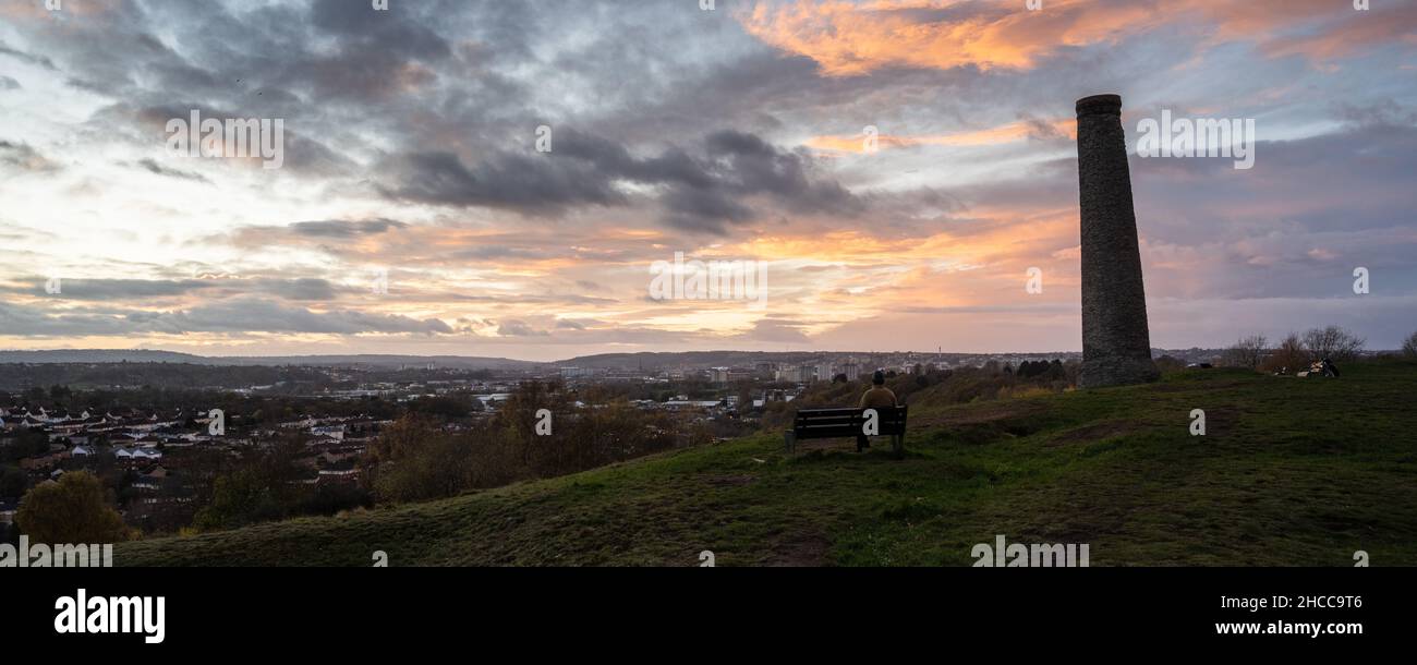 Ein Mann sitzt auf einer Bank und beobachtet den Sonnenuntergang über dem Stadtzentrum von Bristol vom Troopers Hill Park aus. Stockfoto