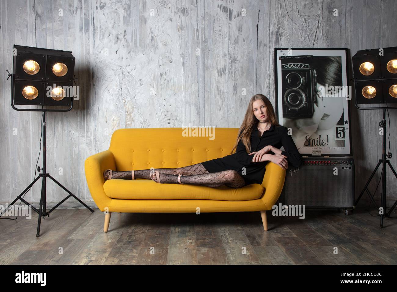 Eine schöne Studioaufnahme eines atemberaubenden blonden professionellen Models, das auf einem gelben Sofa mit einem schwarzen kurzen Kleid, hohen Absätzen und Strumpfhosen sitzt Stockfoto