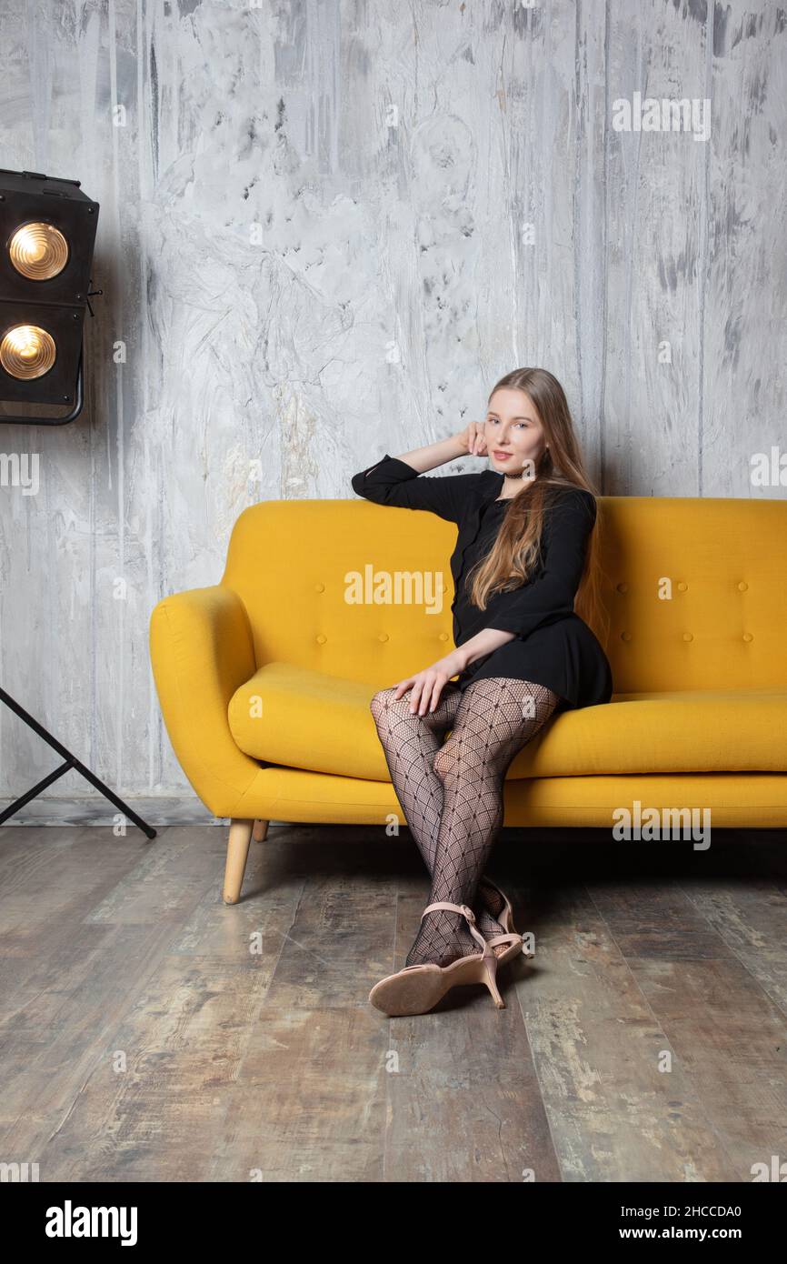 Eine schöne Studioaufnahme eines atemberaubenden blonden professionellen Models, das auf einem gelben Sofa mit einem schwarzen kurzen Kleid, hohen Absätzen und Strumpfhosen sitzt Stockfoto