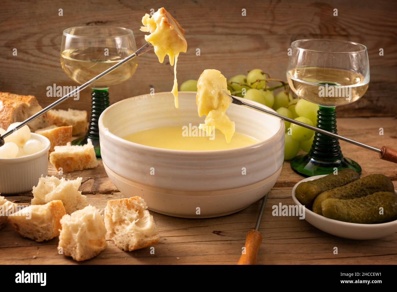 Schweizer Fondue aus geschmolzenem Käse mit Brot auf langen Gabeln, Gurken, Trauben und Wein auf einem rustikalen Holztisch, oft an Feiertagen wie Neujahr serviert Stockfoto