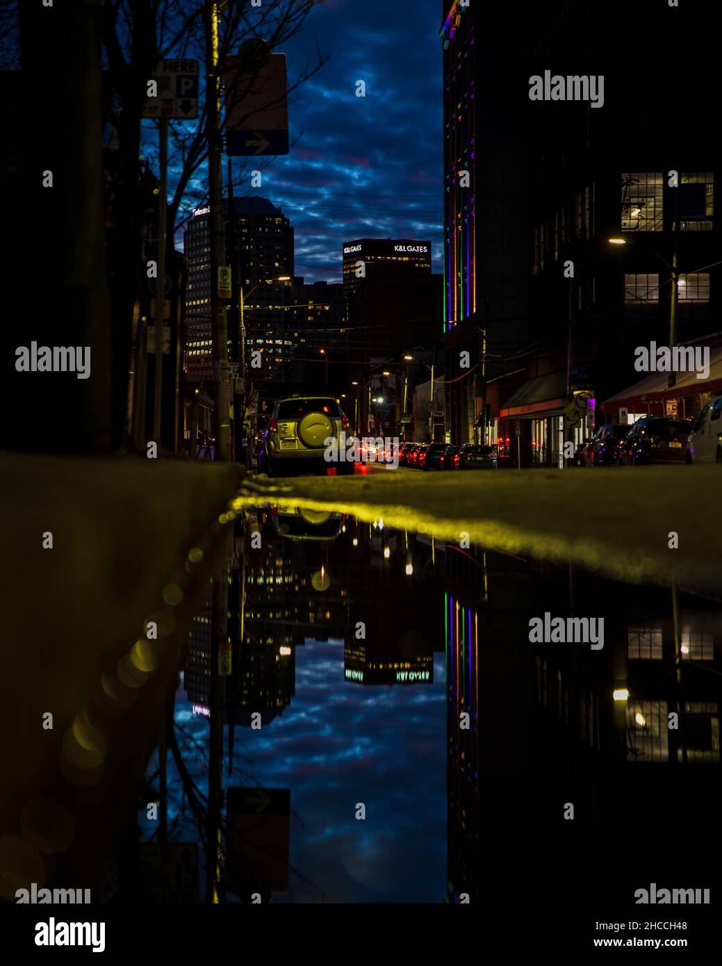 Kleine Pfütze, die hohe moderne Gebäude über die Straße reflektiert, mit geparkten Autos am späten Abend Stockfoto