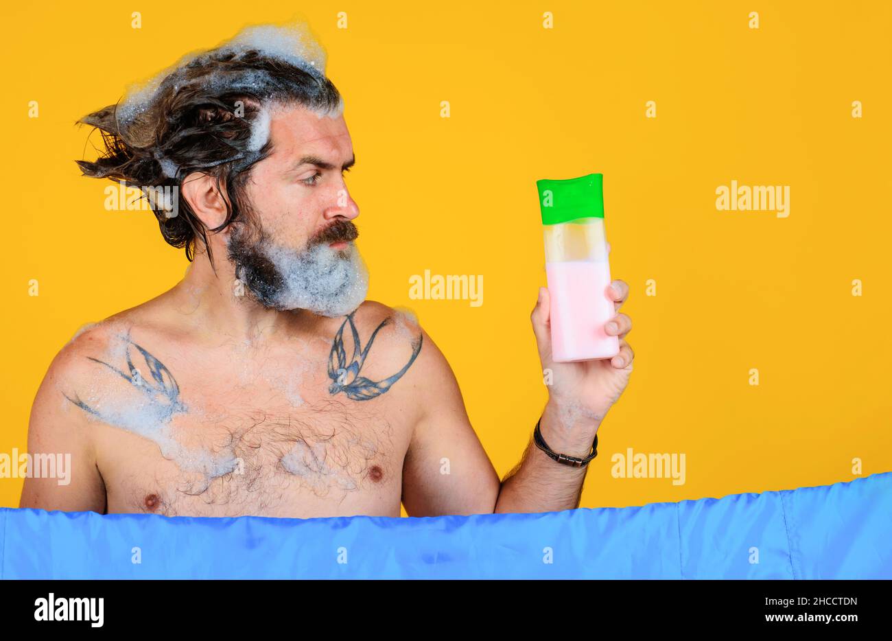 Mann mit Shampoo-Flasche. Bärtiger Mann, der Haare wäscht. Badezeit. Kerl mit Schaum auf dem Kopf. Speicherplatz kopieren. Stockfoto