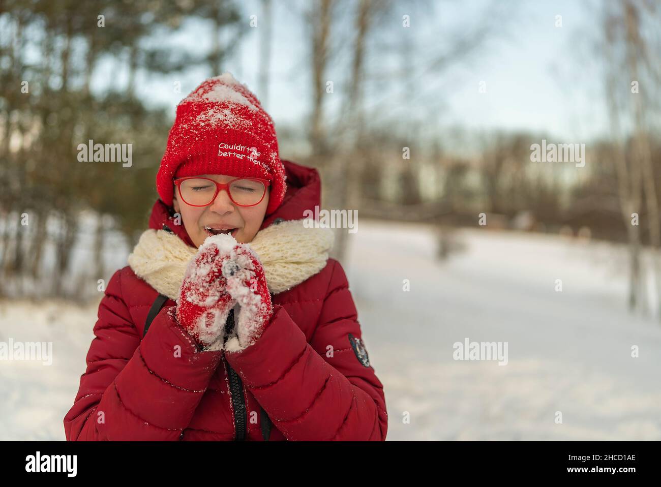 Mädchen in einer roten Jacke und Hut, die Schnee auf dem Hintergrund eines Winterwaldes fressen Stockfoto