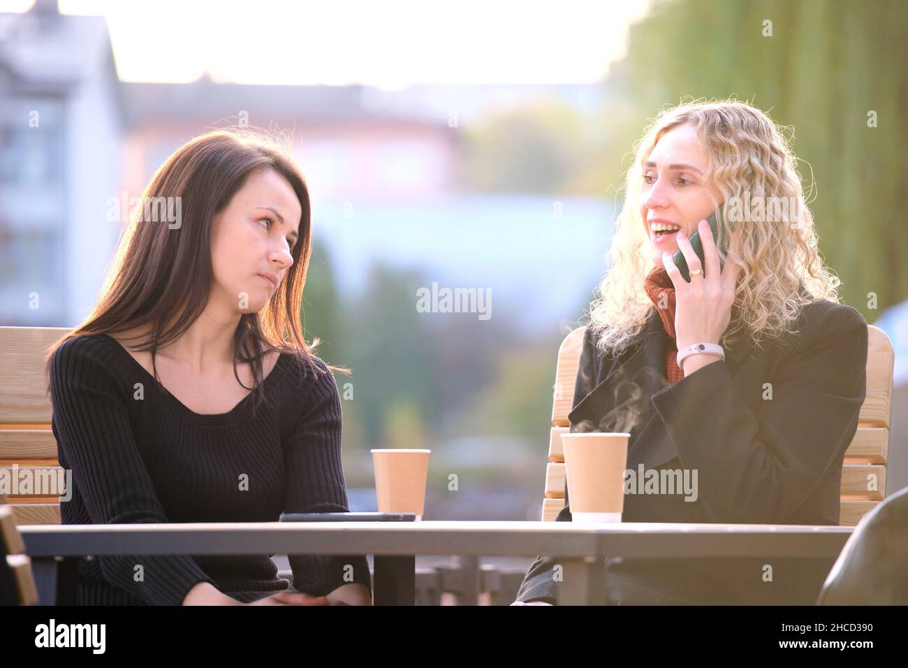 Traurige Frau, die von ihrer Freundin ignoriert wird, die im Straßencafé im Freien sitzt, während sie glücklich auf dem Handy redet und keine Aufmerksamkeit schenkt. Freundschaft Stockfoto