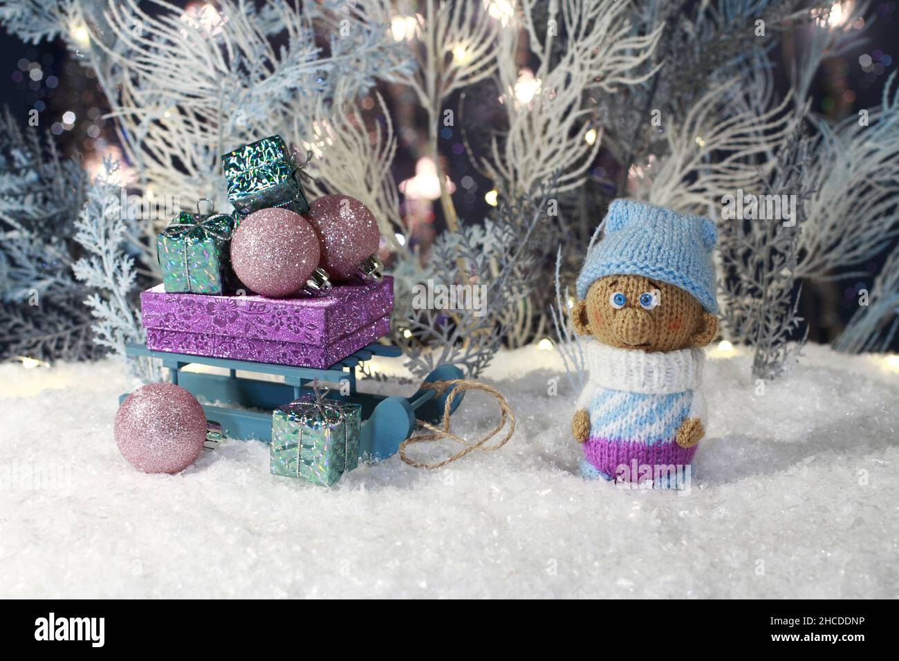 Ein gestrickter Spielzeugknome steht auf dem künstlichen Schnee und ein Spielzeugschlitten mit Geschenken steht neben ihm vor dem Hintergrund eines weißen und blauen Kunstbaums Stockfoto