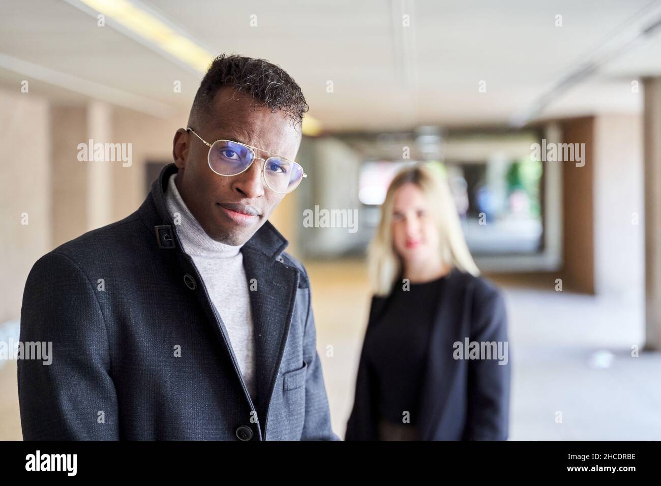 Afroamerikanischer Mann in stilvollem Mantel und Brille, der die Kamera anschaut, während er in einer modernen Passage in der Nähe einer Freundin steht Stockfoto