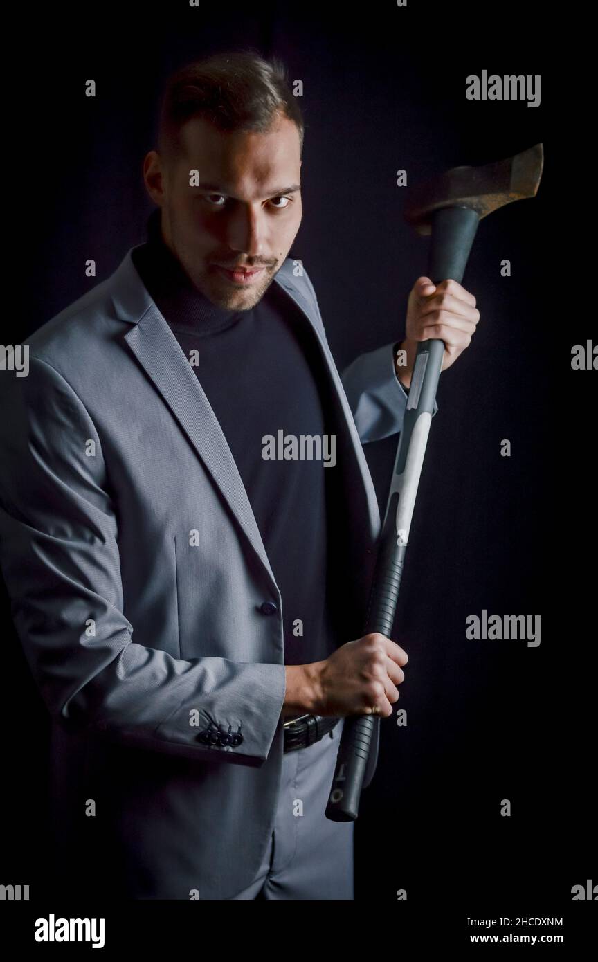 Der junge Geschäftsmann mit grauem Anzug, der eine Axt in der Hand hält, hat ein entschlossenes und bedrohliches Aussehen - ein aggressives Trading-Methodenkonzept Stockfoto