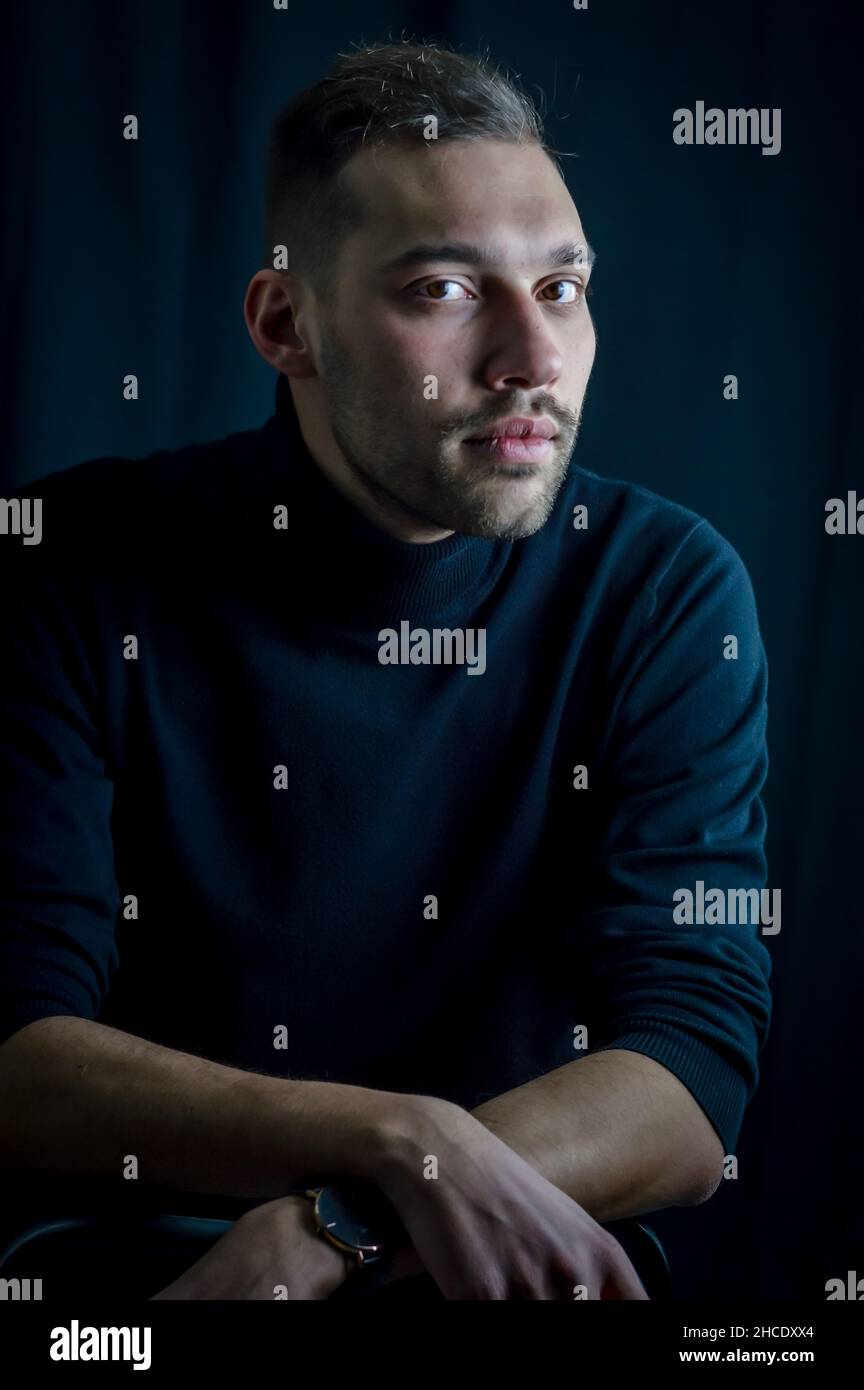 Das Porträt eines hübschen jungen Mannes, der mit gekreuzten Handgelenken sitzt, sieht ernsthaft vor ihm aus Stockfoto