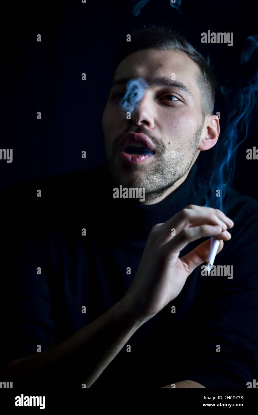 Porträt eines konzentrierten jungen Mannes, der auf schwarzem Hintergrund Rauchringe mit Zigarette macht Stockfoto