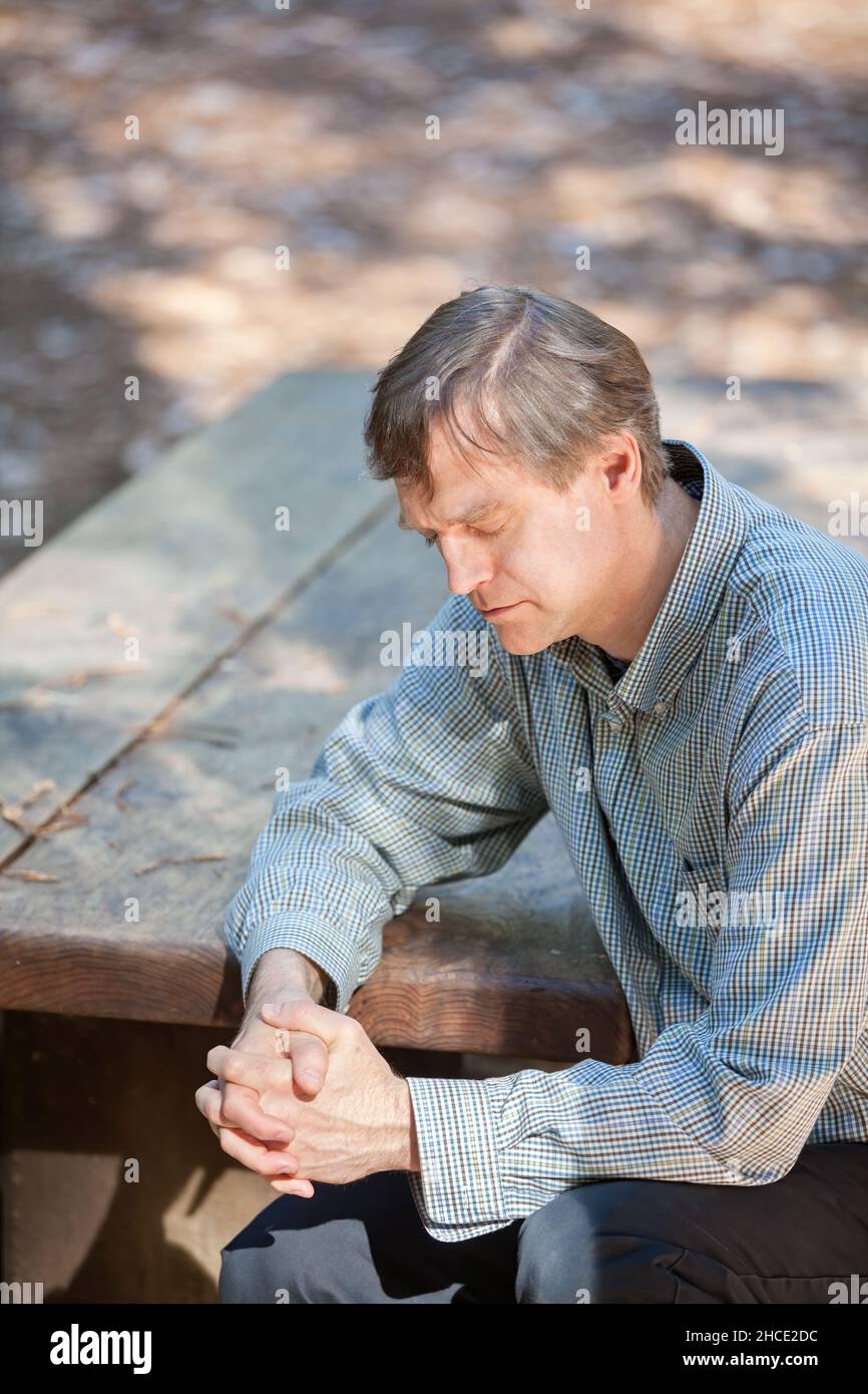 Kaukasischer Mann in den Fünfzigern, der allein am Holztisch sitzt und im Wald betet Stockfoto