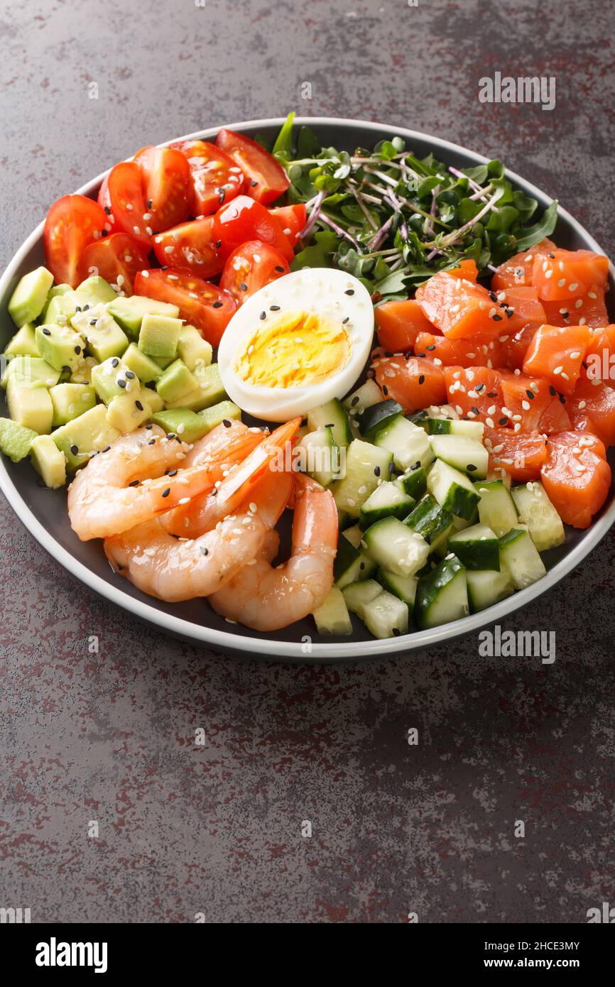 Köstlicher gesunder Salat aus Lachs, Garnelen, Eiern, Avocado, Gurken und Tomaten in Nahaufnahme auf einem Teller auf dem Tisch. Vertikal Stockfoto