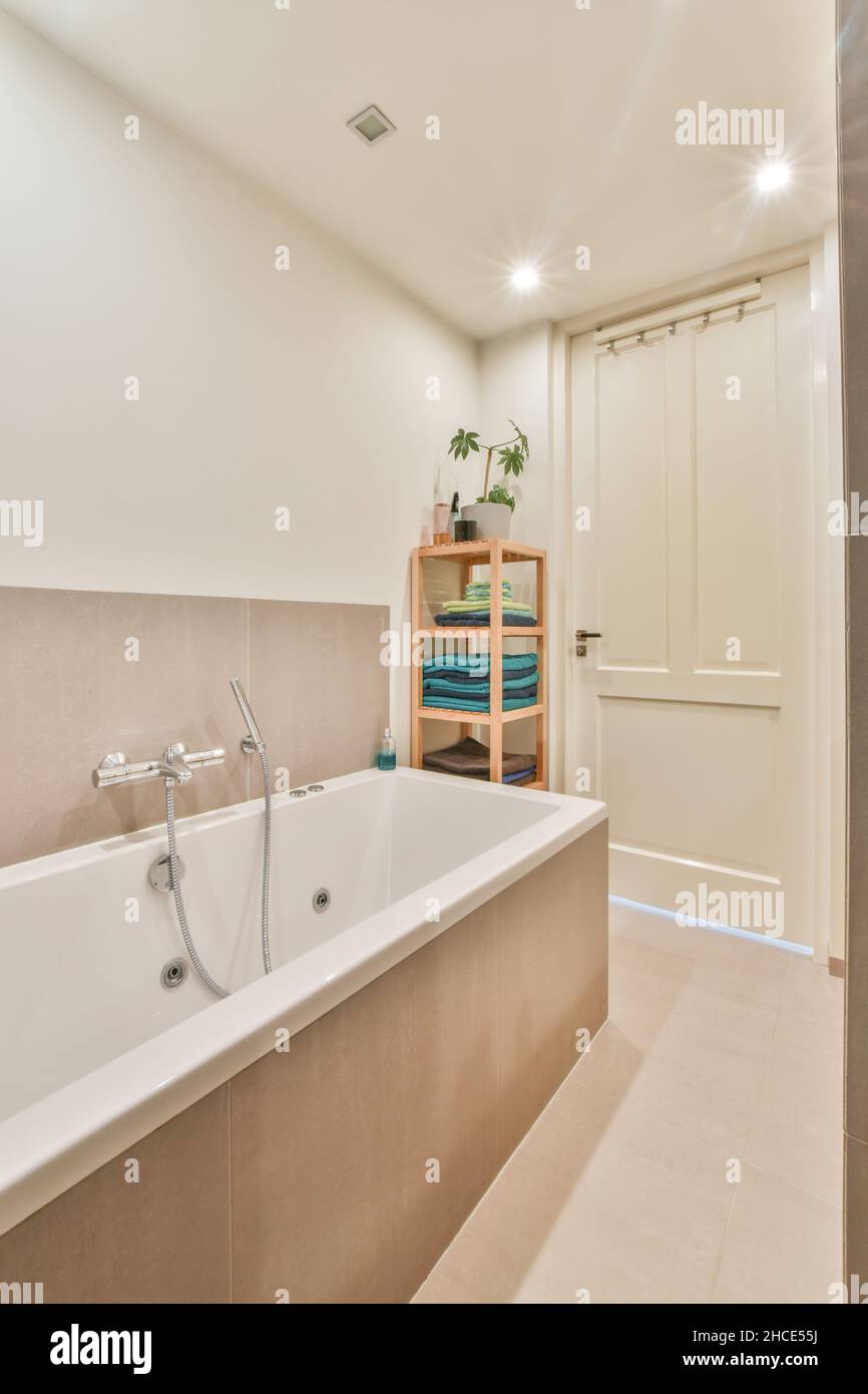 Weiße Badewanne neben einem Regal mit Handtüchern im modernen beleuchteten Badezimmer und im Hintergrund die Zugangstür mit einem Handtuchhalter Stockfoto
