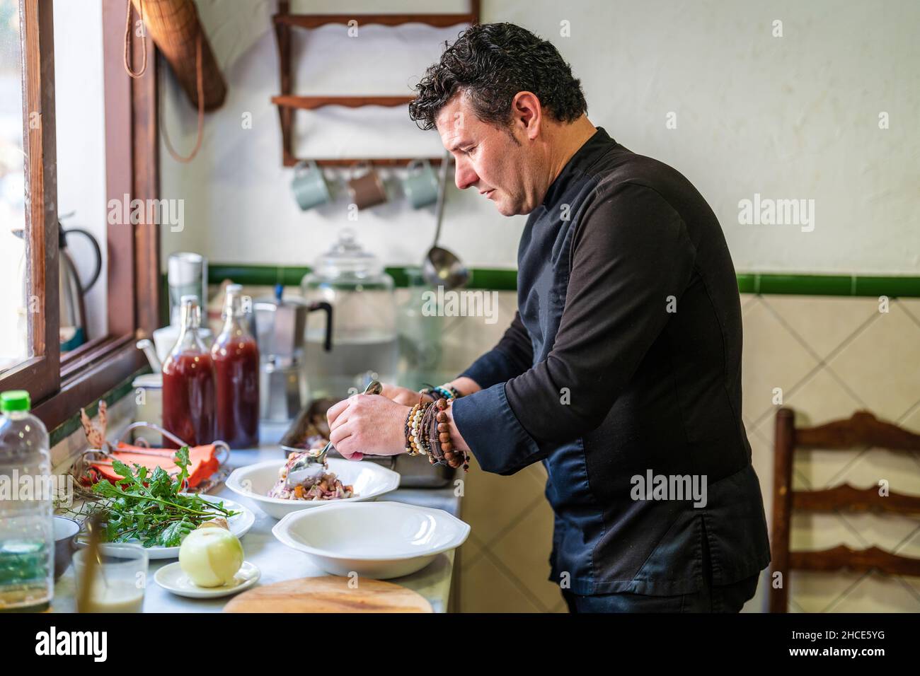 Seitenansicht des männlichen Küchenchefs in schwarzer Uniform, der leckere Speisen auf dem Tisch serviert und verschiedene Zutaten in der Küche enthält Stockfoto