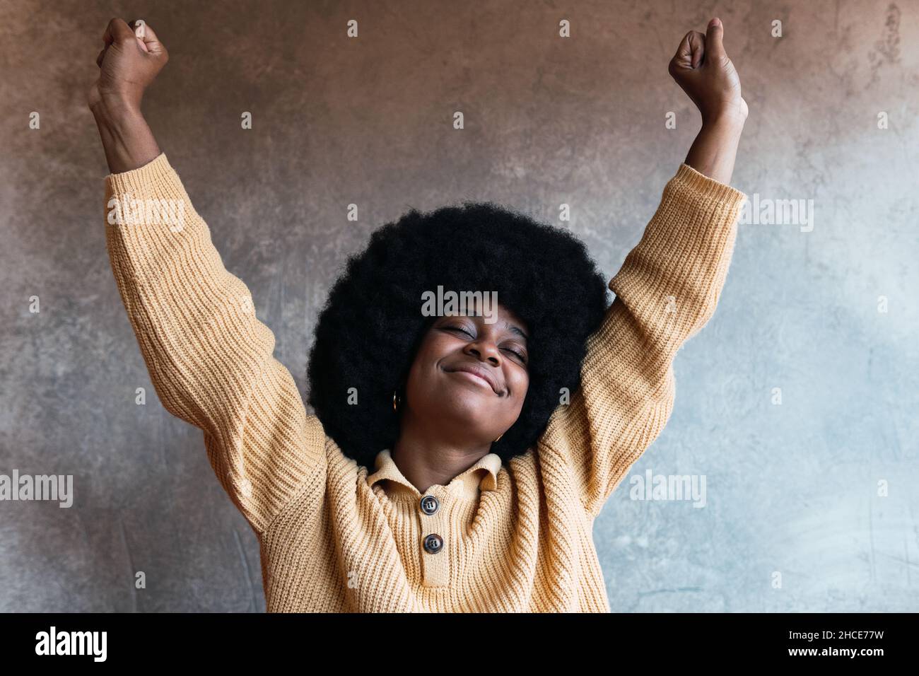 Entzückte afroamerikanische Frau mit Afro-Frisur und geschlossenen Augen, die sich mit erhobenen Armen in Heimkleidung streckte, nachdem sie zu Hause dagegen aufwachte Stockfoto